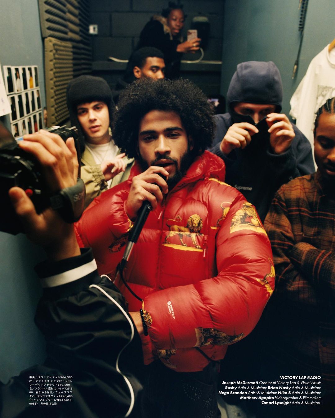 Man in a red puffer jacket holds a microphone, surrounded by a diverse group in a music studio, capturing a candid, vibrant scene.