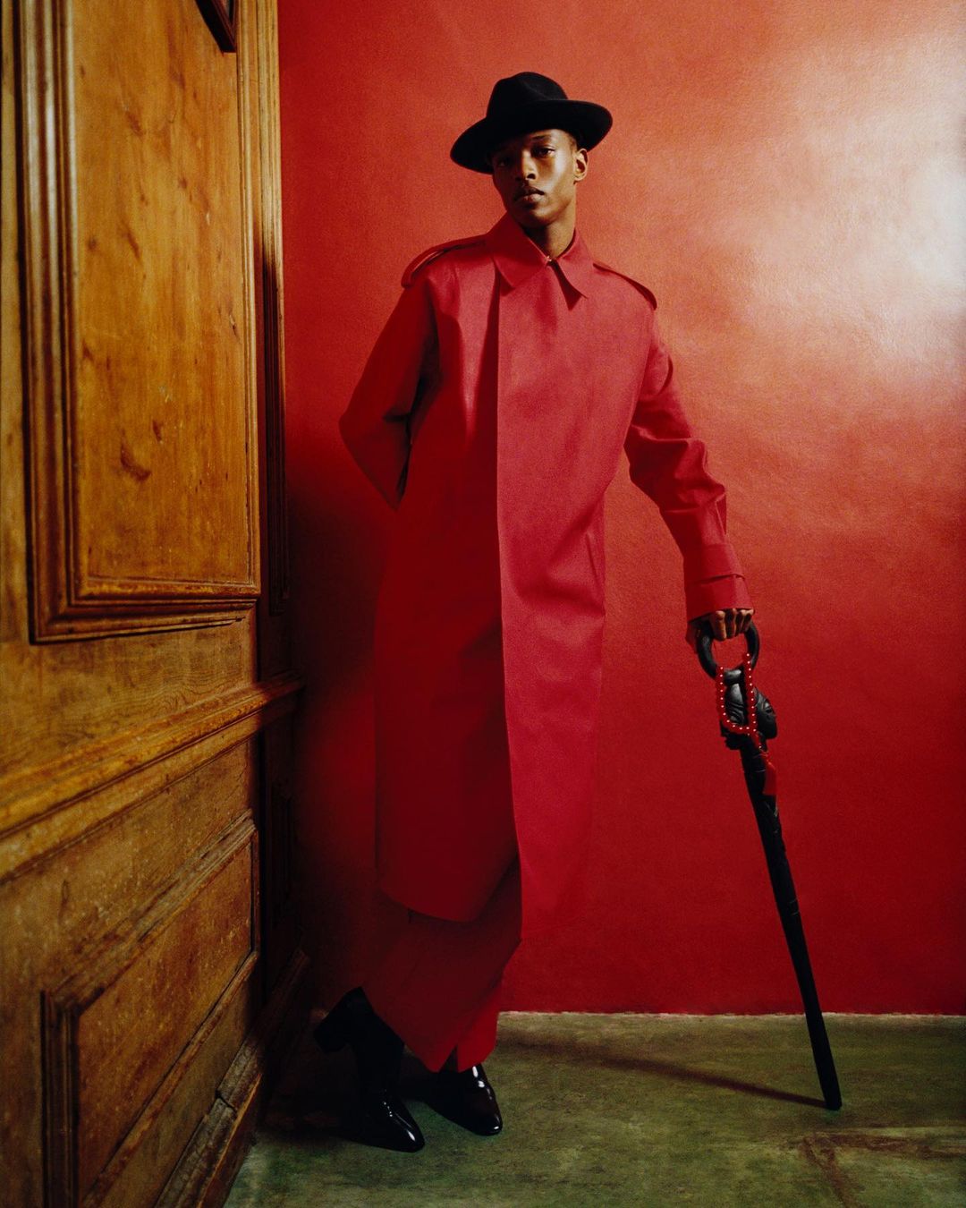 Man in red coat and black hat poses with cane against a red wall next to wooden door. Fashion editorial shot with dramatic lighting.
