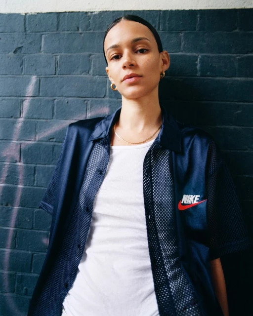 A woman with short brown hair poses indoors against a brick wall in a navy mesh Nike shirt and white top, looking at the camera.