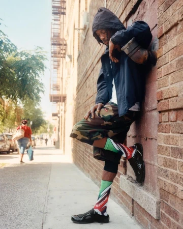 Man wearing a dark hooded jacket and camouflage shorts leaning against a brick wall on a city street