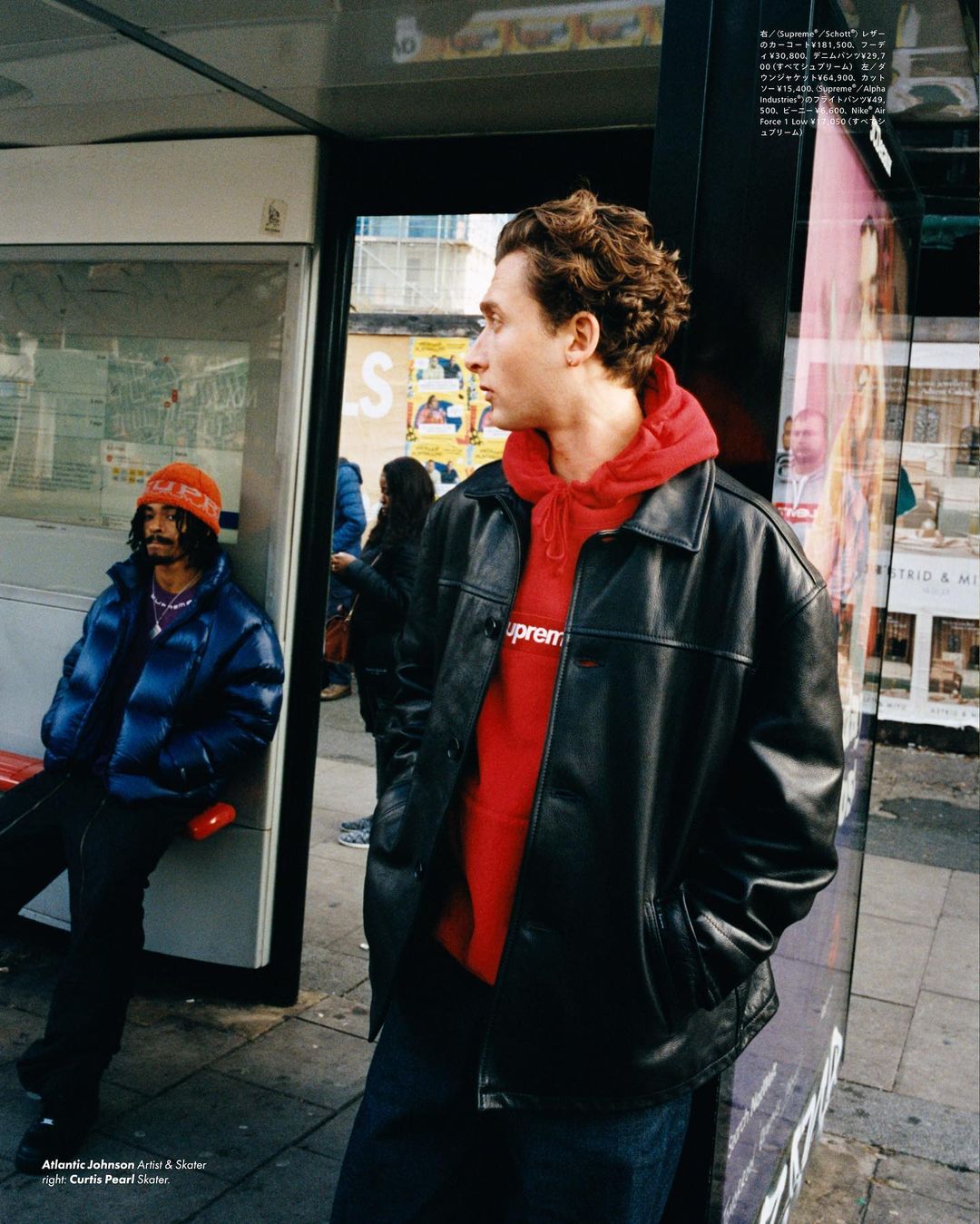 Two men at bus stop: one sitting in a blue puffer jacket and the other standing in a red hoodie and a leather jacket.