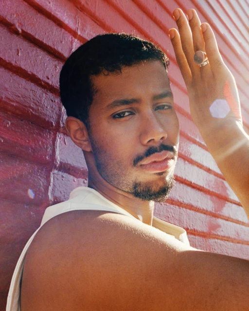 Portrait of a mixed-race man against a reddish-brown brick wall, his hand near his face, adorned with a gold ring, warm light.