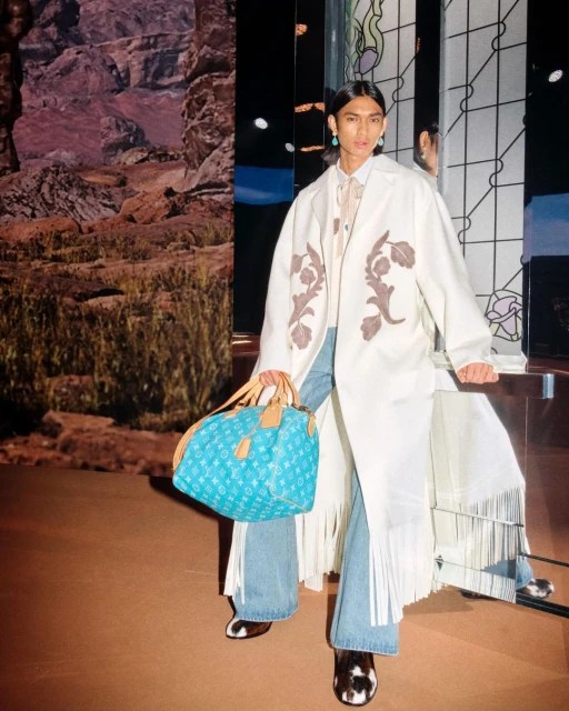 Man poses in long coat, light jeans, and print handbag in front of a mountain scene and patterned glass wall indoors.