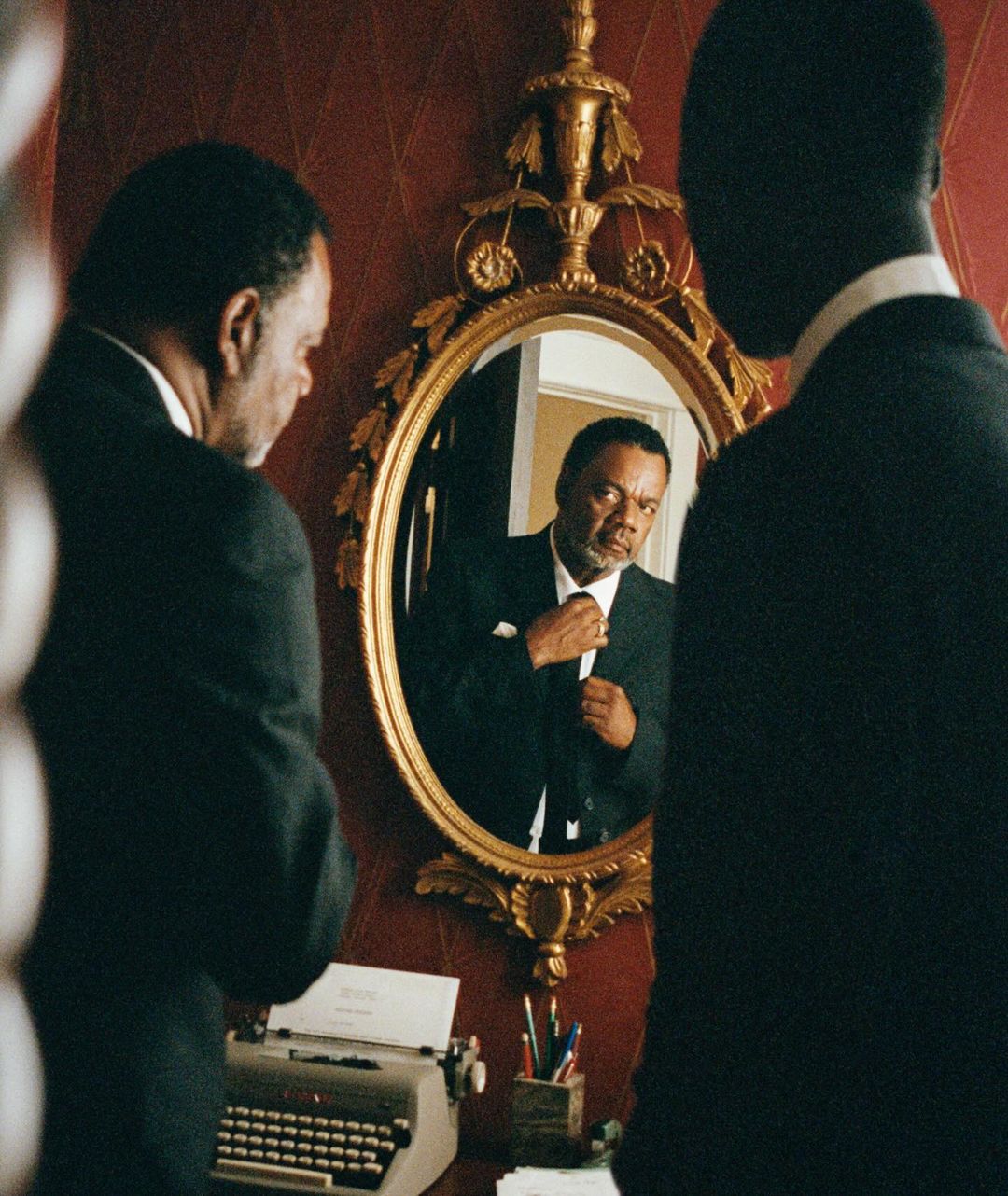 An older Black man in a suit adjusts his tie in a gold-framed oval mirror, vintage typewriter and desk accessories are visible in the setting.
