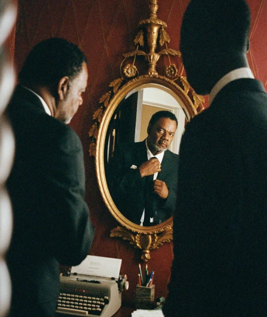 An older Black man in a suit adjusts his tie in a gold-framed oval mirror, vintage typewriter and desk accessories are visible in the setting.