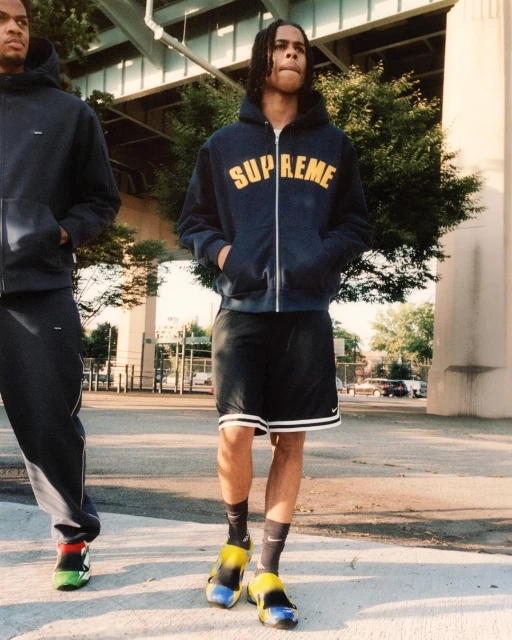 Two men in navy hoodies and athletic shorts walk on a sunny urban street under a bridge, showcasing a casual fashion style.