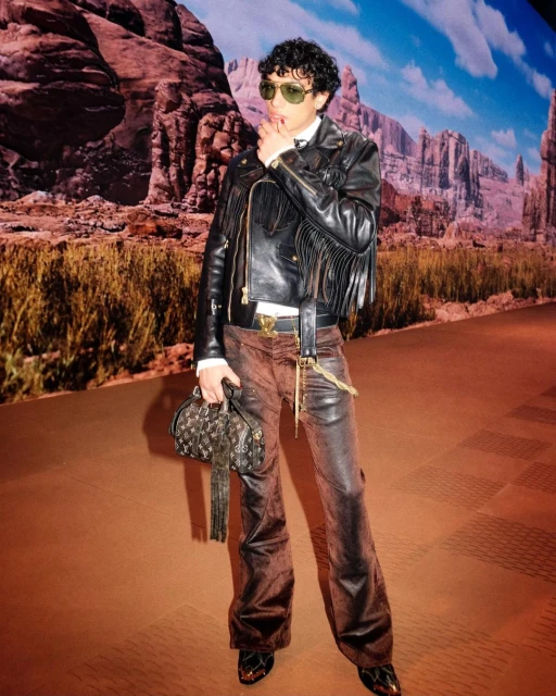A person wearing a black leather fringed jacket, brown pants, and sunglasses poses in front of a Western-themed backdrop. 