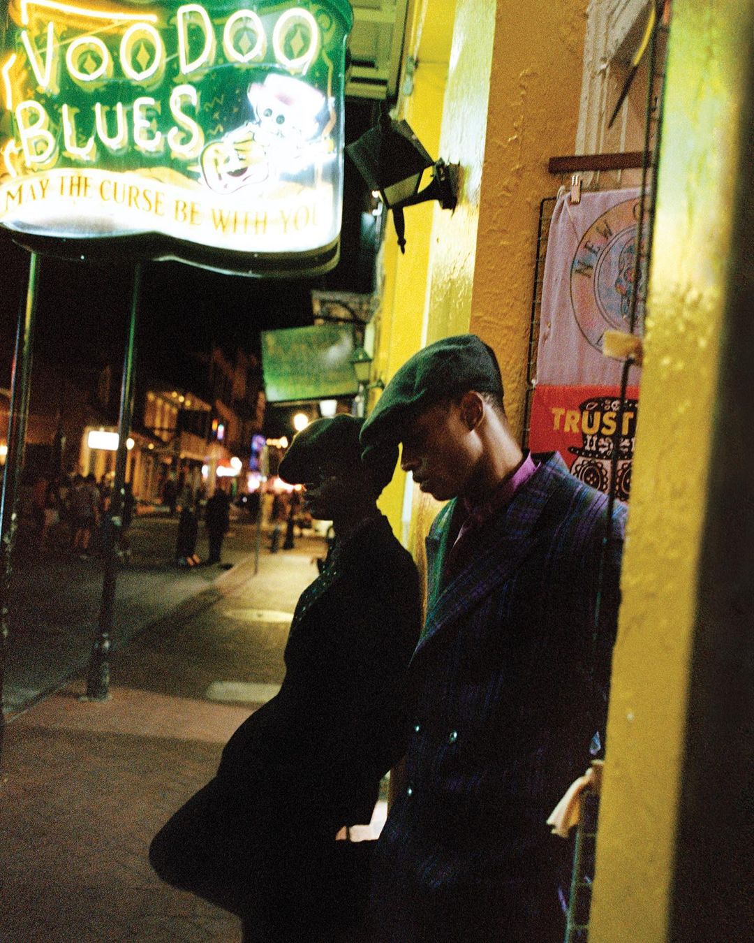 Two men in hats stand on a city street at night, leaning against a yellow-painted wall under a Voodoo Blues sign with streetlights.