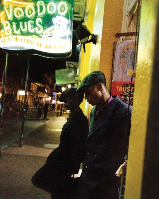 Two men in hats stand on a city street at night, leaning against a yellow-painted wall under a Voodoo Blues sign with streetlights.