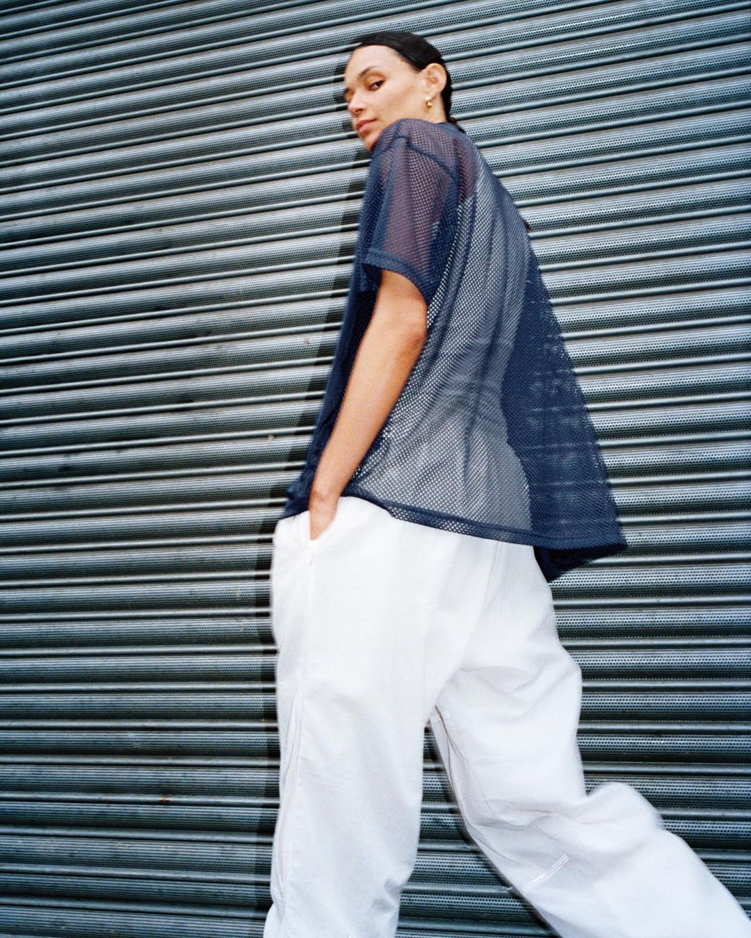 Woman in mesh t-shirt and white pants poses in front of a gray roller shutter. Fashion shot with eye contact and soft lighting.