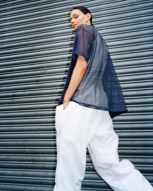 Woman in mesh t-shirt and white pants poses in front of a gray roller shutter. Fashion shot with eye contact and soft lighting.