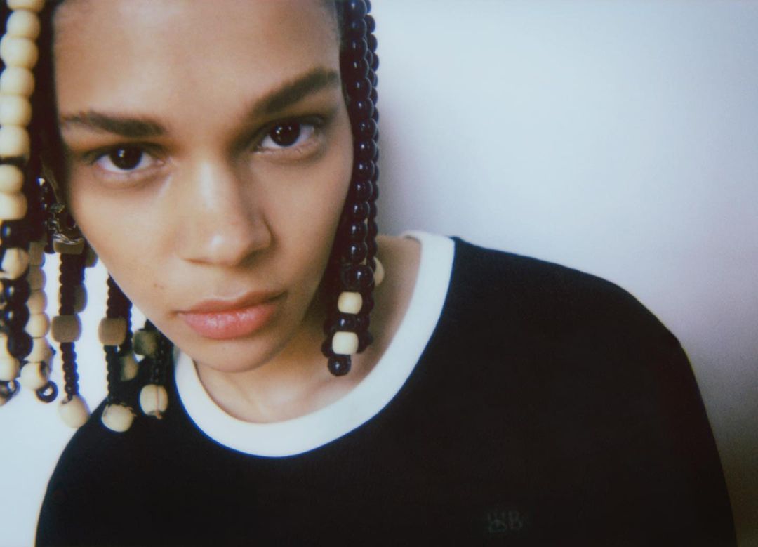 Close-up portrait of a young adult female with beaded braids wearing a black, ringer T-shirt against a neutral white background.