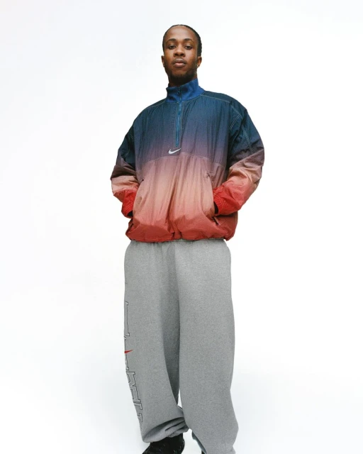 An adult person in a faded ombre windbreaker and baggy sweatpants stands in a photo studio with a white backdrop, hands in pockets.