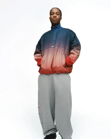 An adult person in a faded ombre windbreaker and baggy sweatpants stands in a photo studio with a white backdrop, hands in pockets.