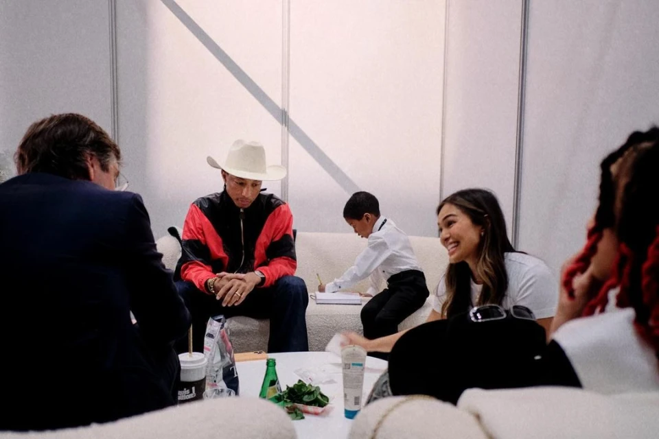 Indoor scene featuring Pharrell Williams in a cowboy hat, seated on a couch alongside a child drawing, and two women, casual setting.
