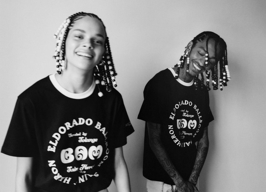 A smiling woman and man wear graphic t-shirts in a black and white indoor studio portrait. Their hair is styled with beads.