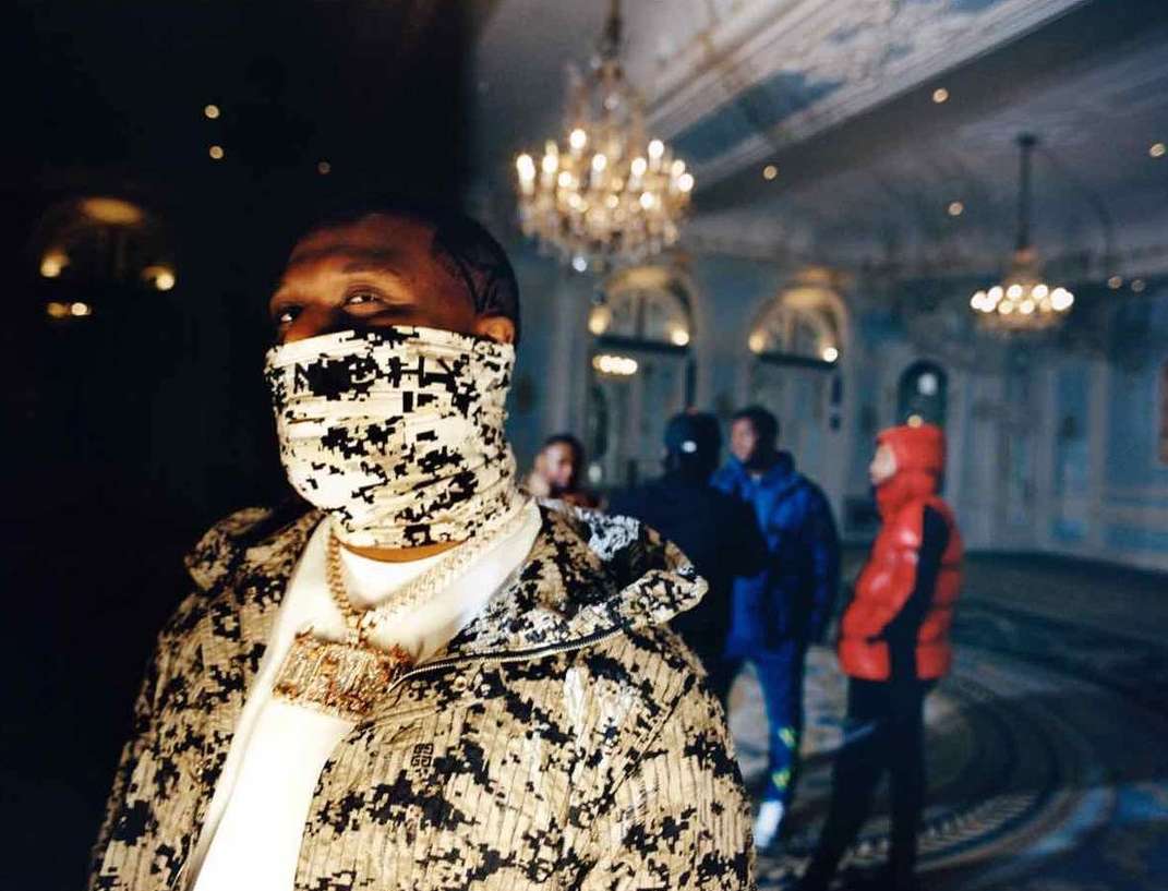 Man in patterned jacket and face covering stands in opulent room with group; chandelier, arched doorways, and decorative floor.