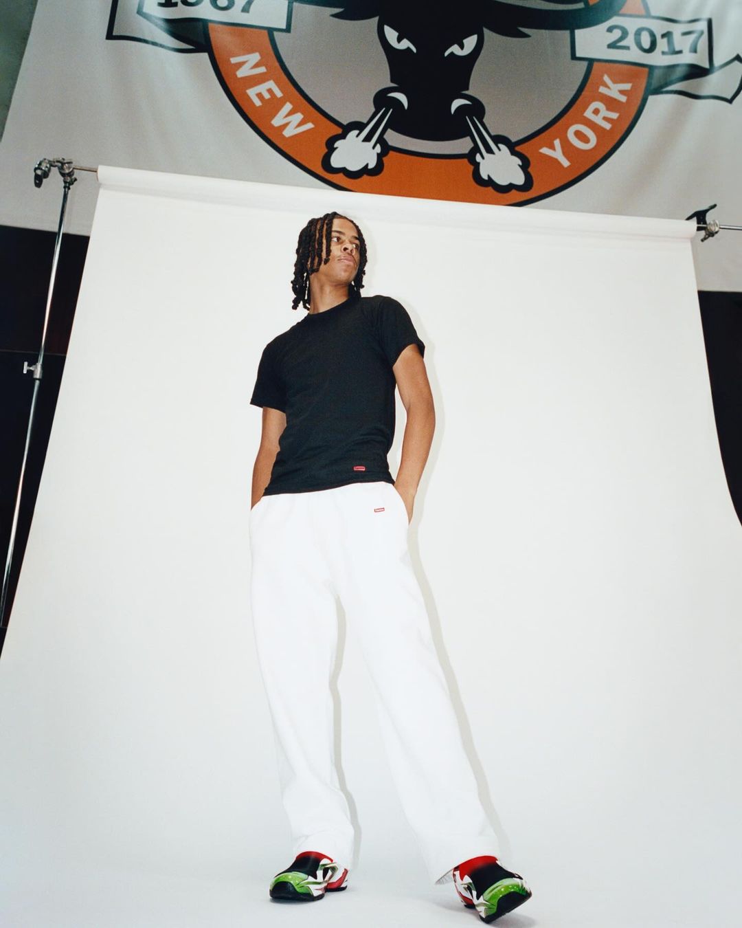 Man in black t-shirt and white pants stands in a photo studio with a sports logo banner backdrop, illuminated by studio lighting.