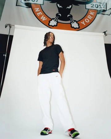 Man in black t-shirt and white pants stands in a photo studio with a sports logo banner backdrop, illuminated by studio lighting.