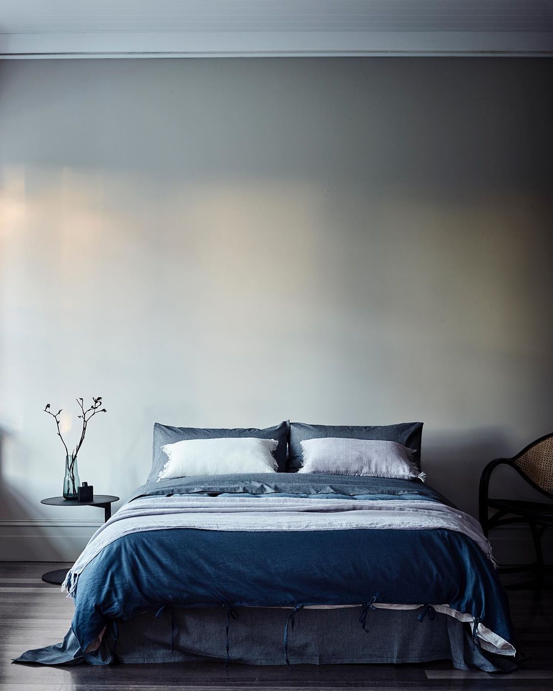 Bedroom interior with a made bed featuring a dark blue duvet, white pillows, and a side table with a glass vase and branches.