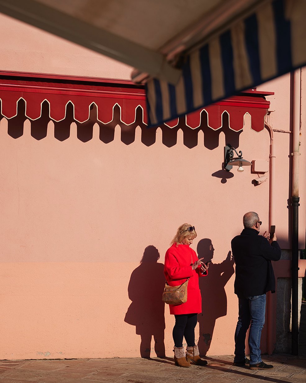 Two people using mobile phones near a pink wall with red awning, casting shadows on a sunny day. Architectural details visible.