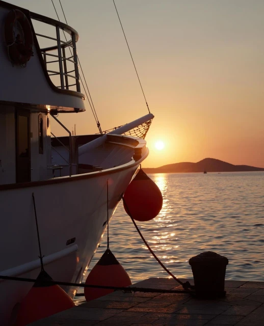 Boat docked at sunset, featuring an eye-level shot of a white boat with red buoys against a sunset backdrop with calm ocean waters.
