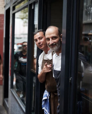 Two chefs in aprons smile out from the window of a restaurant, one pointing towards the camera in an inviting gesture.