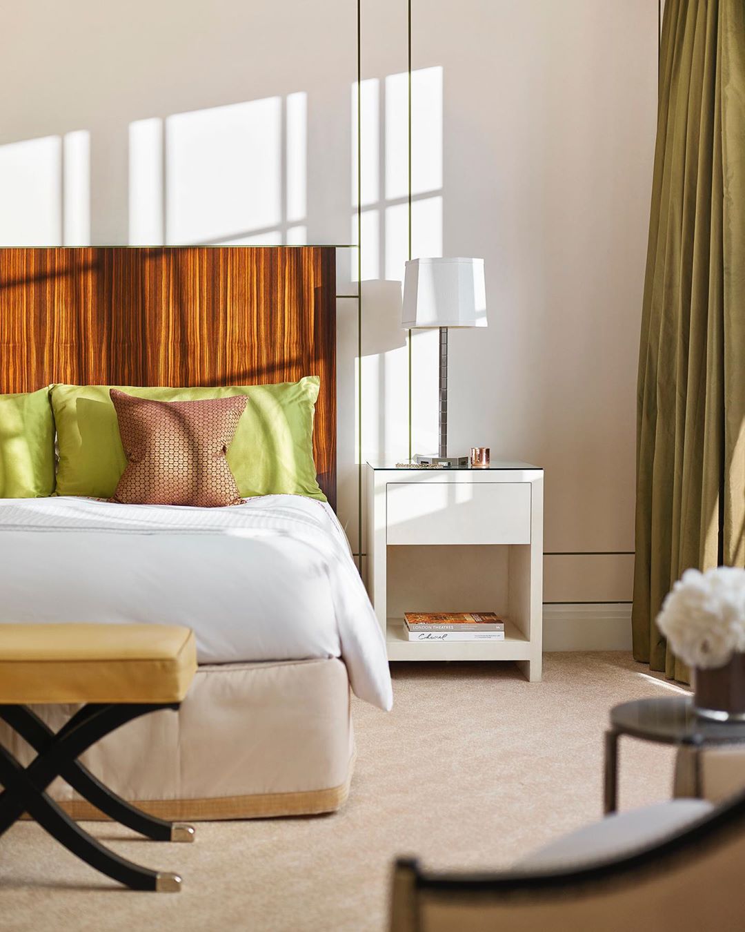 Interior shot of a neutral-toned bedroom features a wooden headboard, pillows, side table with a lamp, and an upholstered bench.