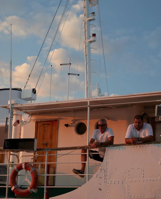 Two adult men on a vintage boat under a blue sky with some clouds, the sun is setting, casting warm hues on the scene.