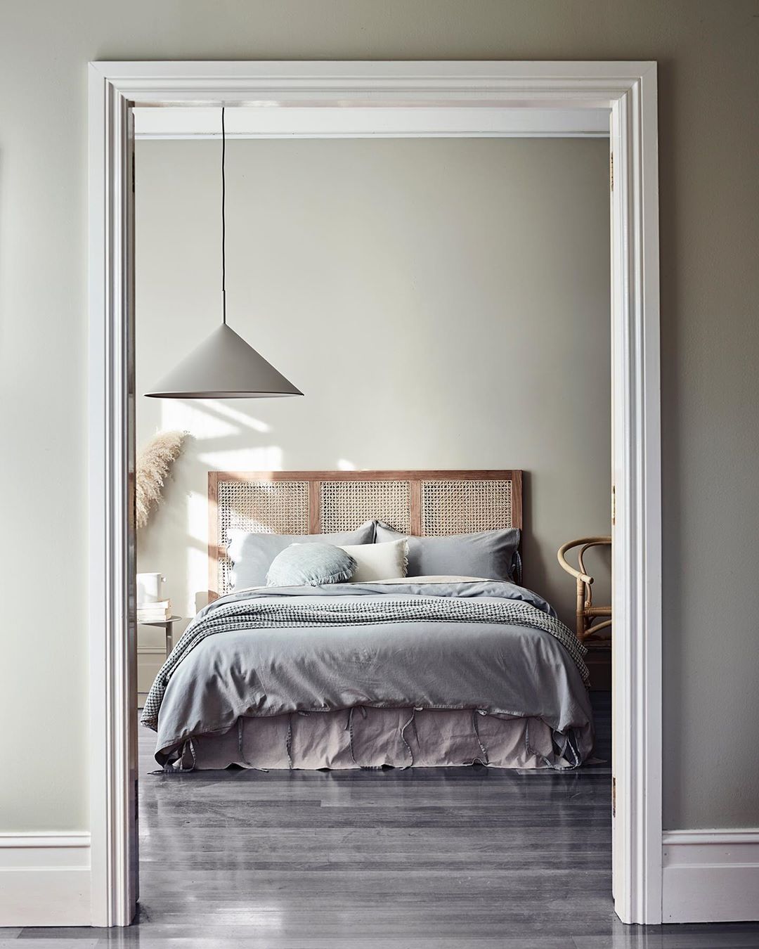 Bedroom interior featuring a bed with a rattan headboard, grey bedding, a hanging lamp, and neutral wall colors, viewed through doorway.