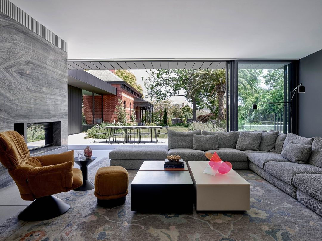 Living room interior with grey L-shaped sofa, tan armchair, fireplace, coffee table, and outdoor view through large sliding glass doors.