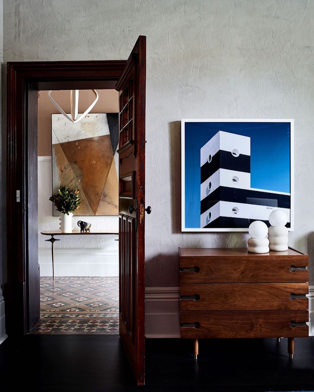 Home interior featuring a wooden door ajar, opening to a tiled floor and an abstract art piece with a wooden dresser and a modern building art piece.