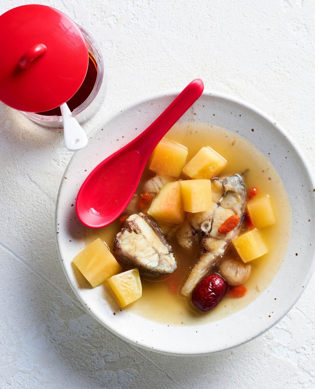 Overhead shot of fish soup with cubed potatoes, red dates, and goji berries in a speckled ceramic bowl alongside a red spoon.