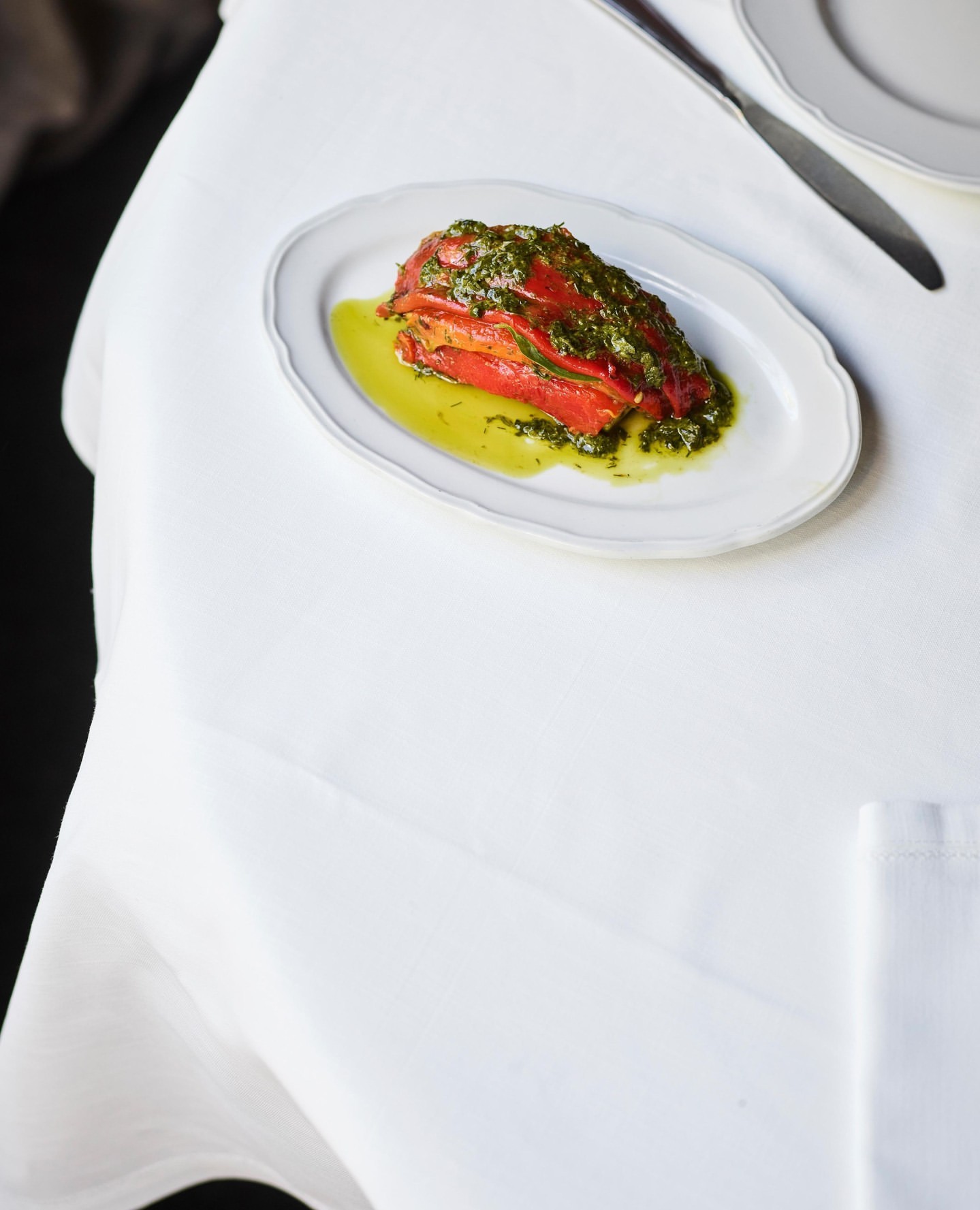 Overhead shot of roasted red peppers topped with green sauce on an oval plate set on a white tablecloth with silverware.