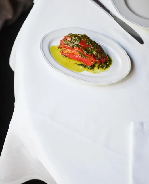 Overhead shot of roasted red peppers topped with green sauce on an oval plate set on a white tablecloth with silverware.