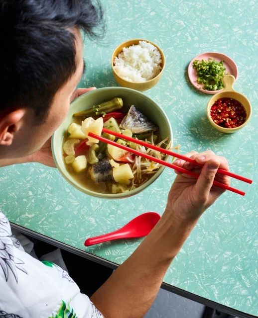 Person eating fish soup with vegetables and rice using chopsticks at a table with sauces and a spoon, food photography.