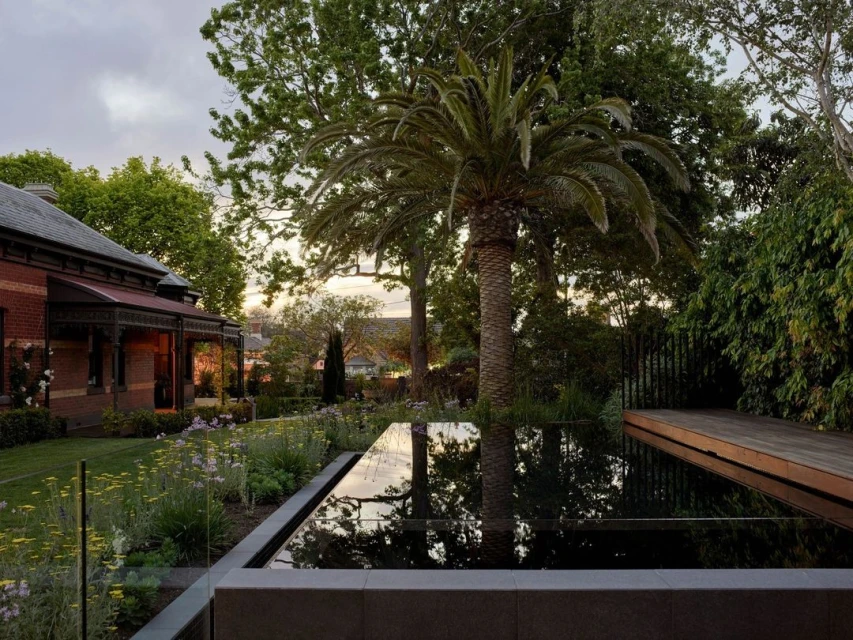 Exterior view of a contemporary garden with a reflecting pool, palm tree, lawn, flowers, and a brick building at dusk.