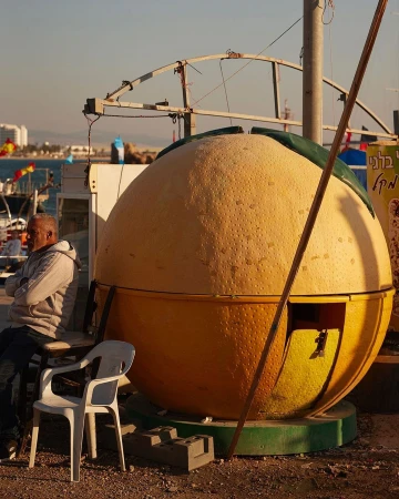 Man seated beside a giant orange shaped kiosk outdoors under natural warm sunlight with a white plastic chair