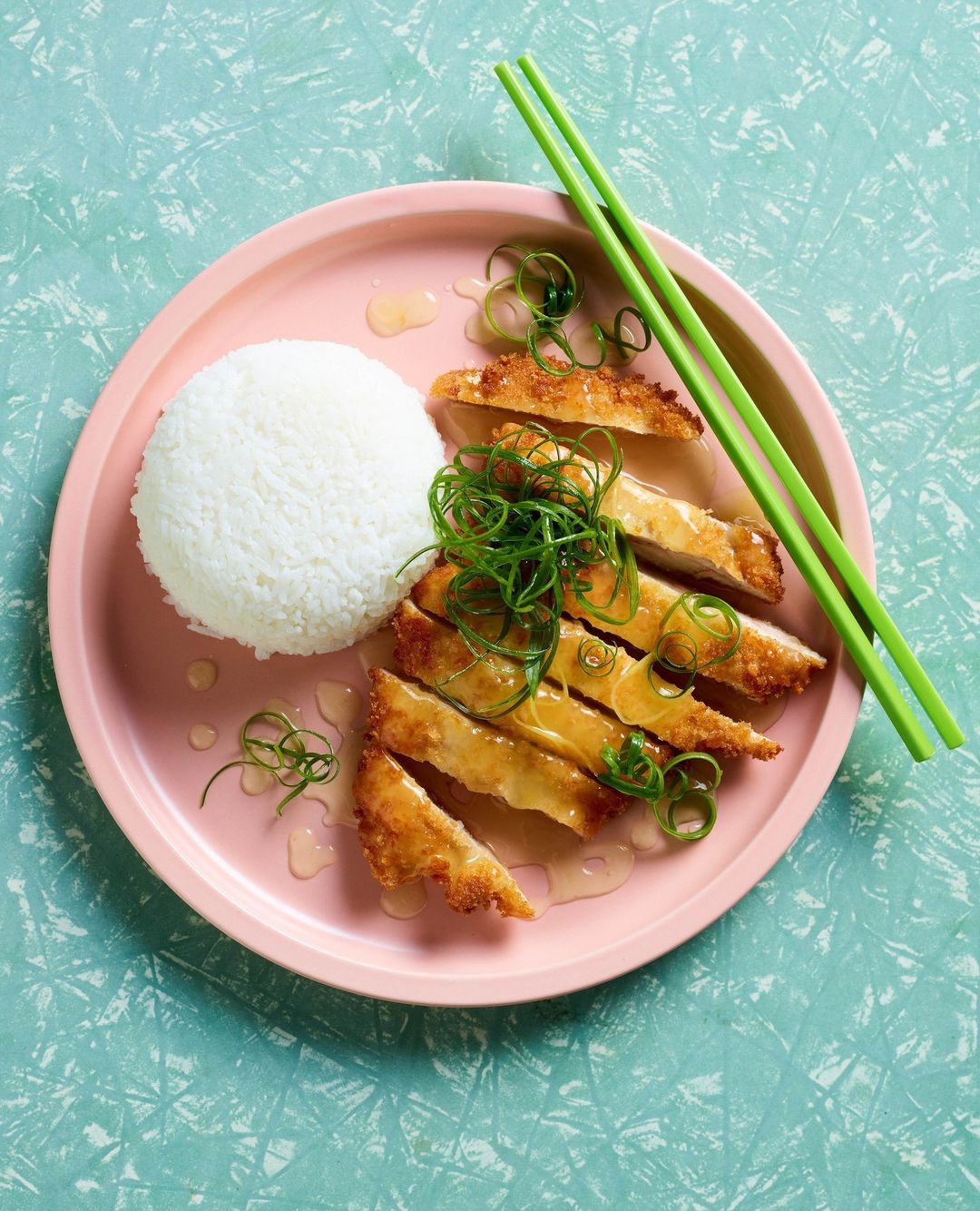 Overhead shot of sliced, fried chicken with white rice and green onions on a pink plate, accompanied by green chopsticks on a teal surface.