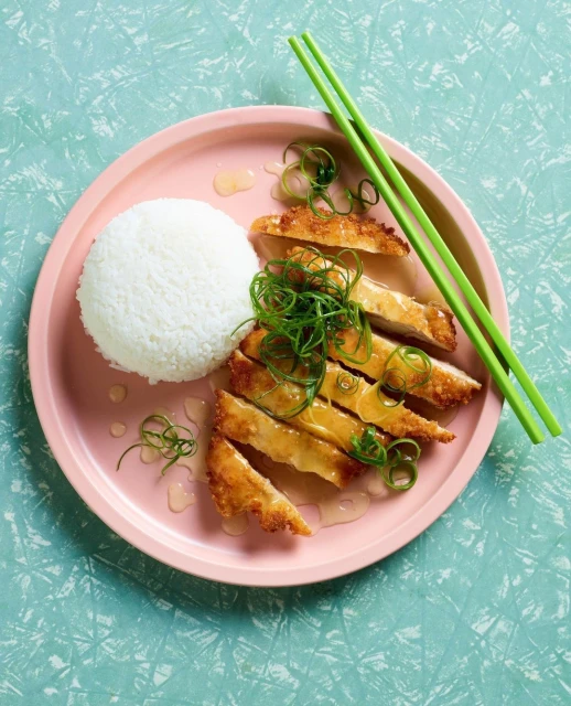Overhead shot of sliced, fried chicken with white rice and green onions on a pink plate, accompanied by green chopsticks on a teal surface.