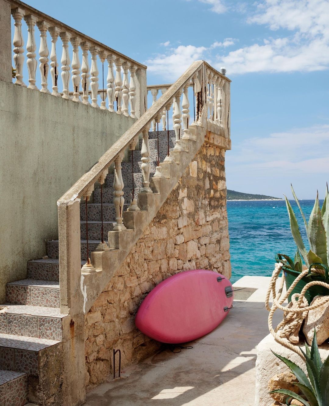 Outdoor stairway with stone balustrade leads to a pink surfboard propped against a stone wall, with the blue sea and sky in the background.