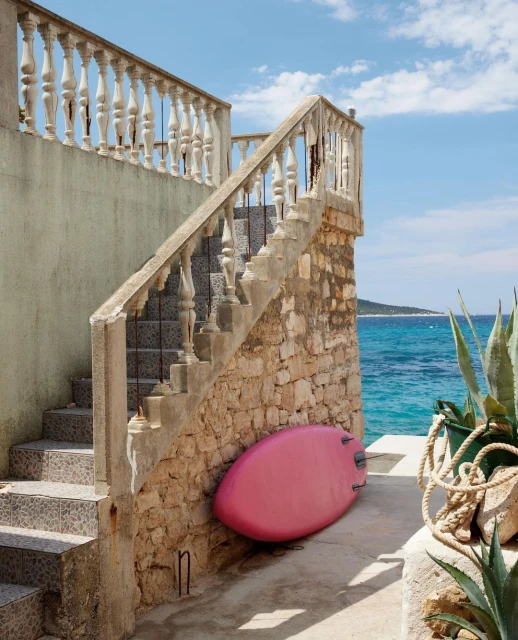 Outdoor stairway with stone balustrade leads to a pink surfboard propped against a stone wall, with the blue sea and sky in the background.