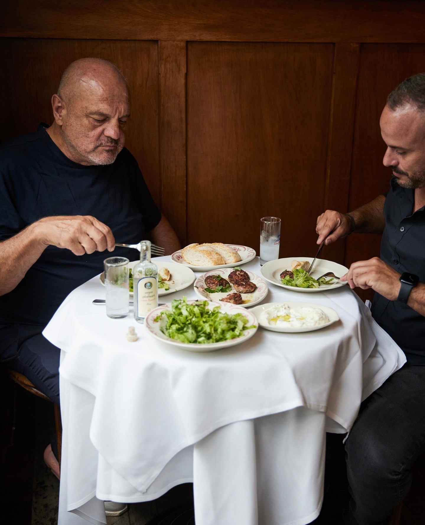 Two men seated at a small round table sharing a meal with plates of food and drinks in a wood paneled restaurant interior.