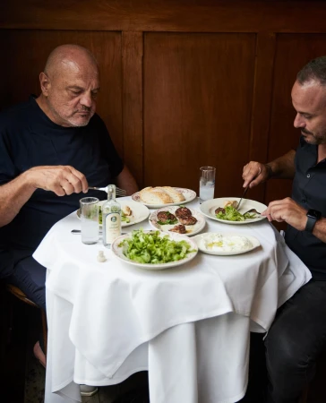 Two men seated at a small round table sharing a meal with plates of food and drinks in a wood paneled restaurant interior.