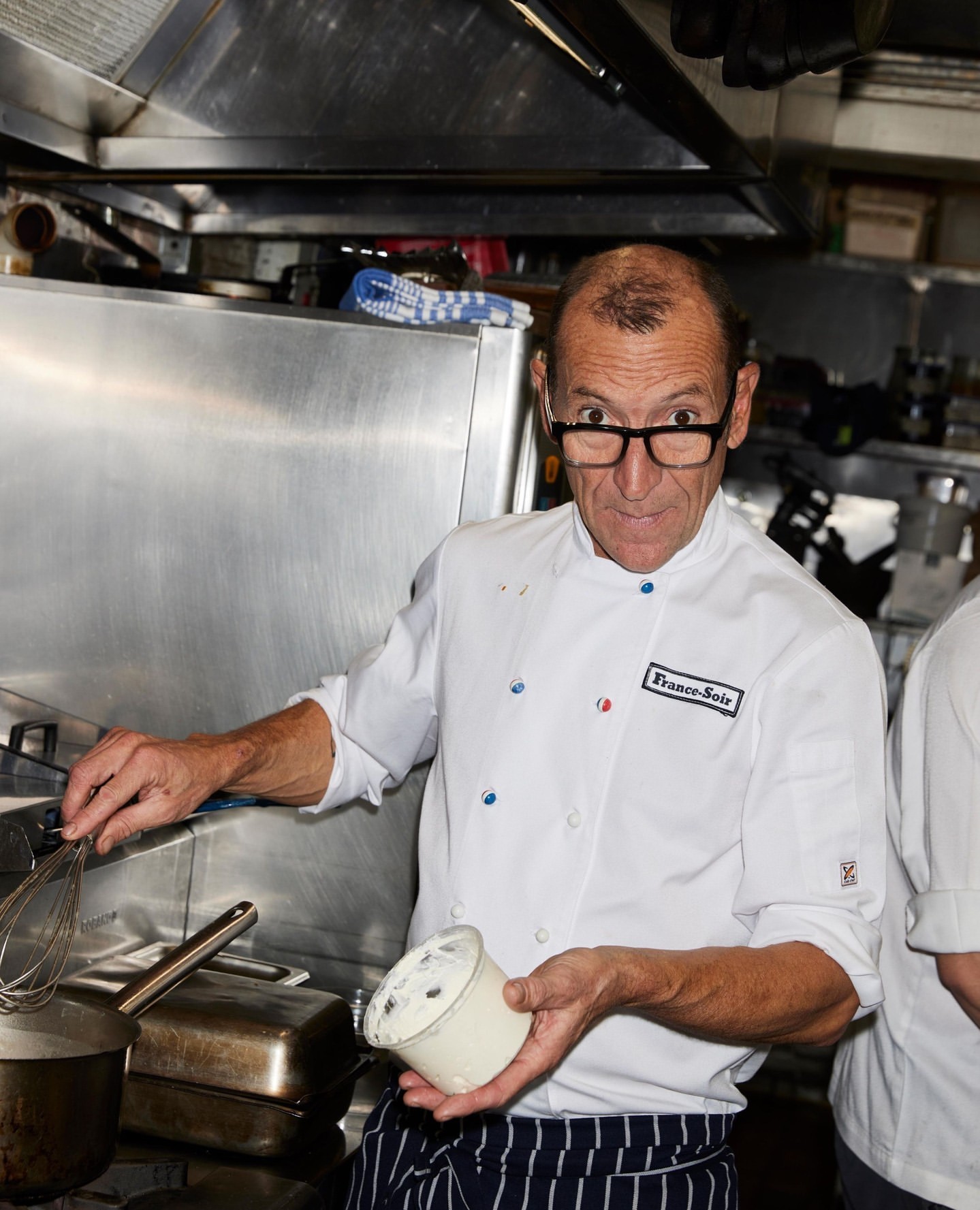 Adult man in chef whites holds a bowl with a whisk in a professional kitchen with stainless steel appliances and cookware.