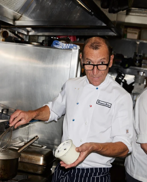 Adult man in chef whites holds a bowl with a whisk in a professional kitchen with stainless steel appliances and cookware.