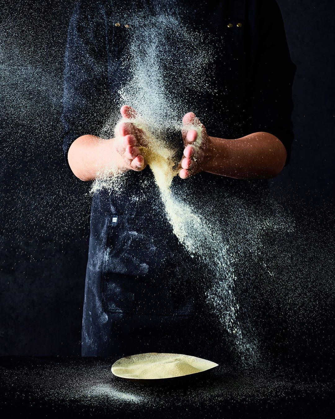 Hands toss flour in a dark studio. The fine powder hangs in the air above a wooden plate covered in flour, creating a dynamic visual.