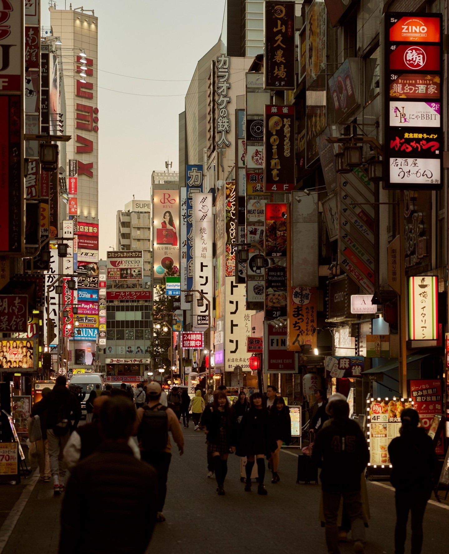 Busy urban street scene in Japan at dusk, with people walking amidst towering buildings covered in illuminated signage.