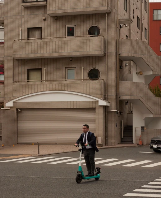 Asian adult male commutes on an electric scooter in an urban setting with a modern building facade along a city street.