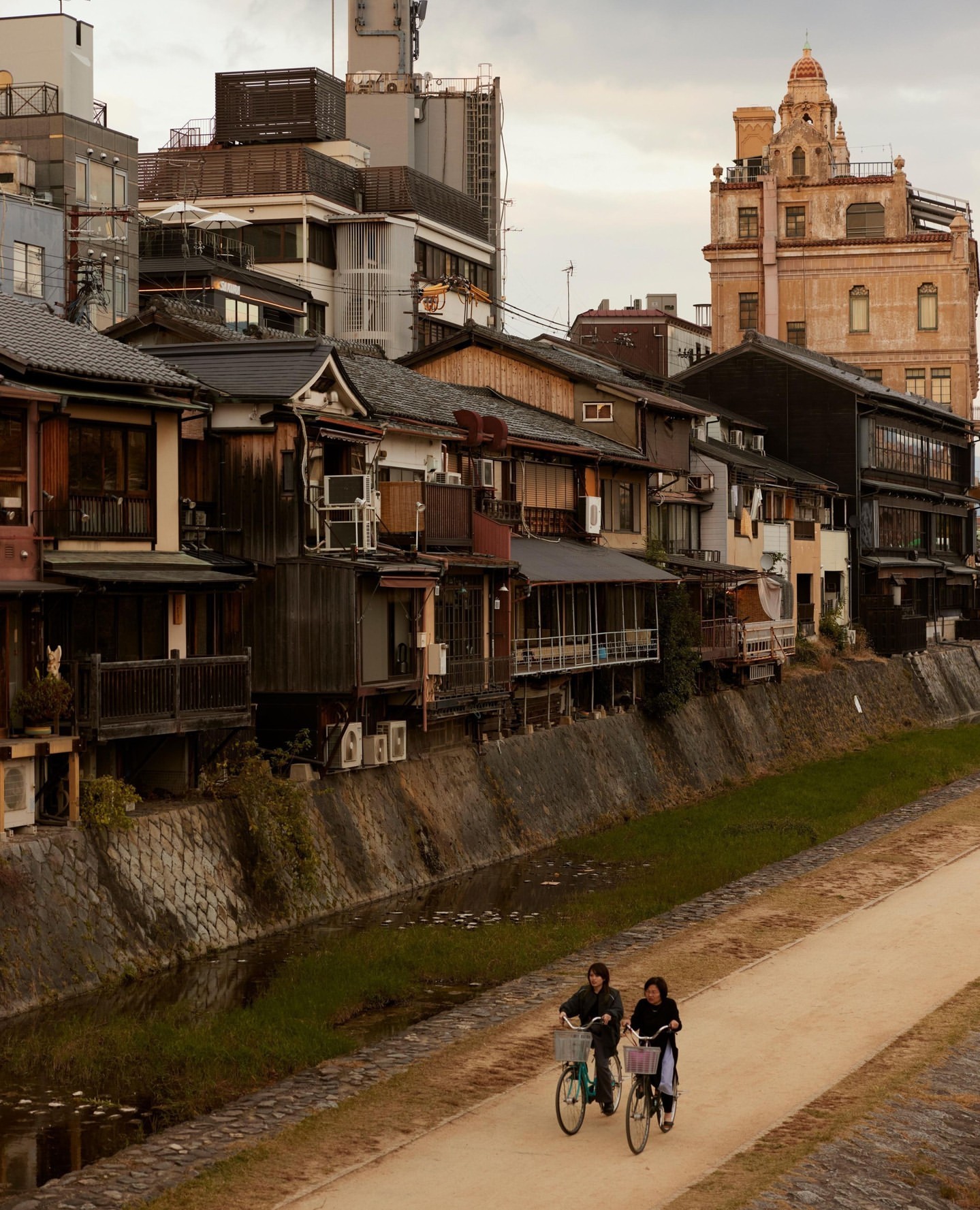 Two people ride bicycles on a path beside a canal in an urban area with traditional Japanese buildings and modern structures in the background.